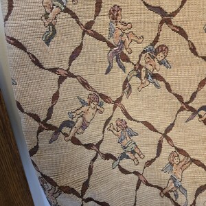 May include: Beige tote bag with a woven pattern featuring cherubs and brown ribbon-like accents. The bag has a rectangular shape and two handles. The cherubs are depicted in various poses with wings and flowing garments.