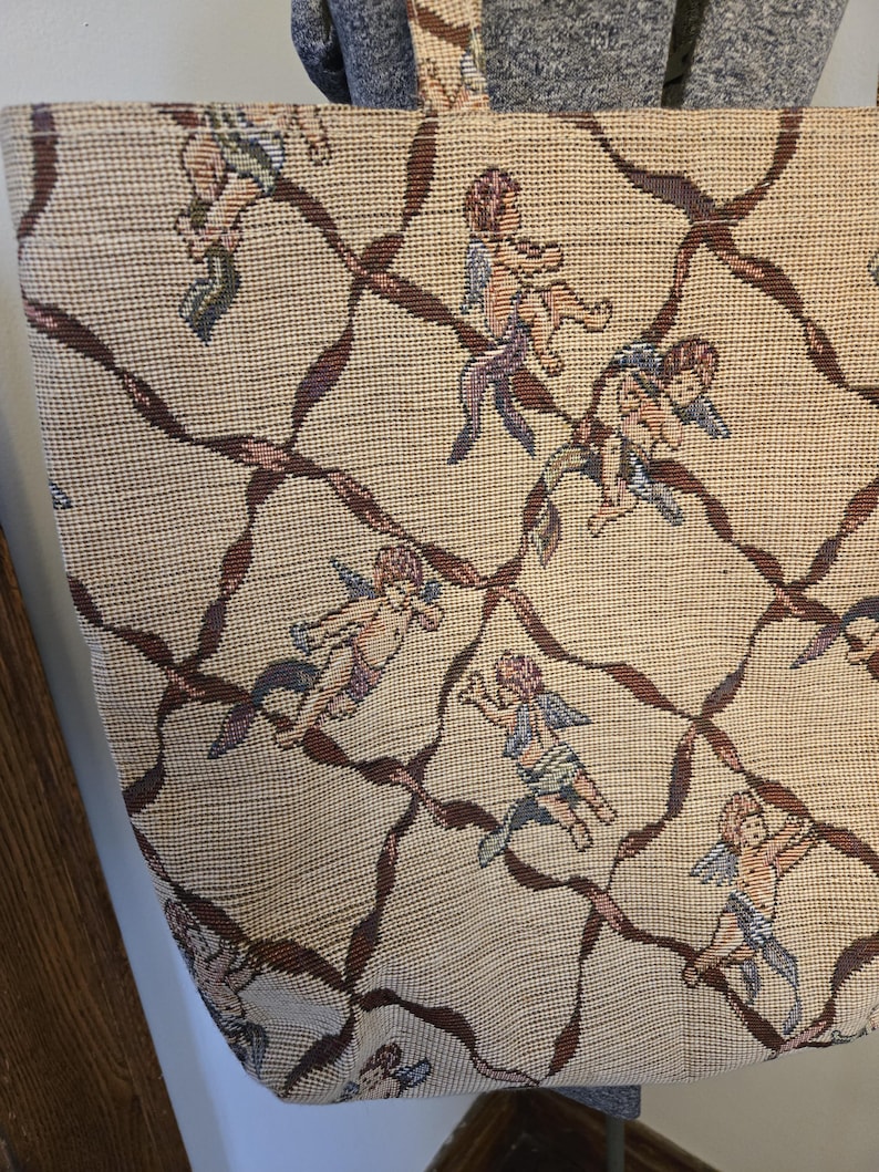 May include: Beige tote bag with a woven pattern featuring cherubs and brown ribbon-like accents. The bag has a rectangular shape and two handles. The cherubs are depicted in various poses with wings and flowing garments.