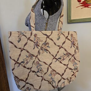 May include: Beige tote bag with a repeating pattern of cherubs and brown ribbon-like lines. The bag has matching handles and is displayed on a mannequin. The bag's design is reminiscent of vintage tapestries.