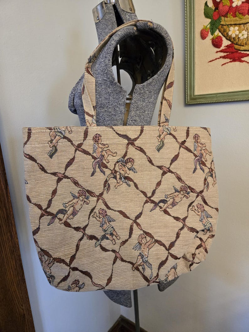 May include: Beige tote bag with a repeating pattern of cherubs and brown ribbon-like lines. The bag has matching handles and is displayed on a mannequin. The bag's design is reminiscent of vintage tapestries.