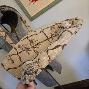 May include: Beige tote bag with a repeating pattern of cherubs and brown vines. The bag has a matching strap and is made of a woven fabric. The bag is held up in the air.