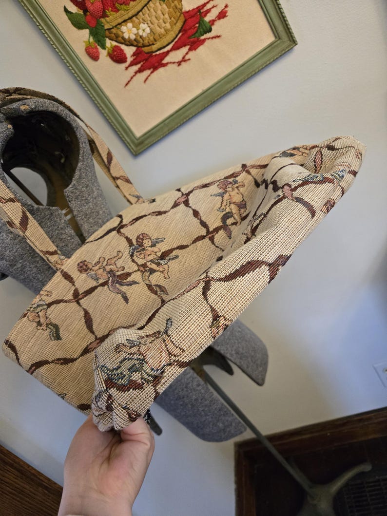 May include: Beige tote bag with a repeating pattern of cherubs and brown vines. The bag has a matching strap and is made of a woven fabric. The bag is held up in the air.