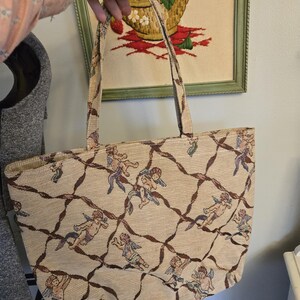 May include: Beige tote bag with a repeating pattern of cherubs and brown lattice design. The bag has two handles and a rectangular shape. The background shows a framed embroidery of strawberries in a basket.