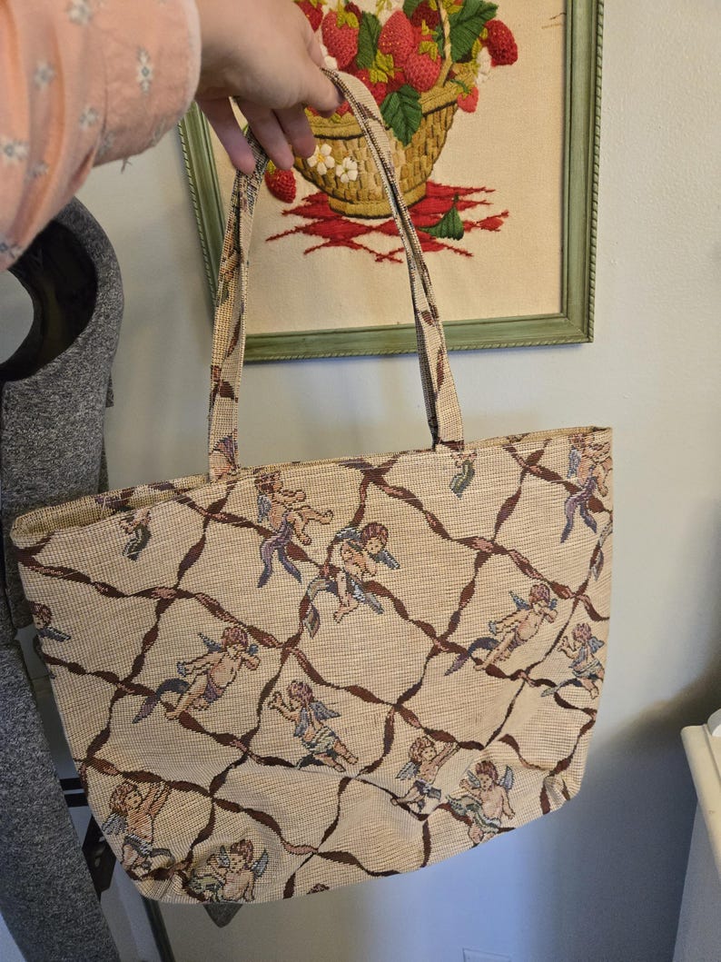 May include: Beige tote bag with a repeating pattern of cherubs and brown lattice design. The bag has two handles and a rectangular shape. The background shows a framed embroidery of strawberries in a basket.