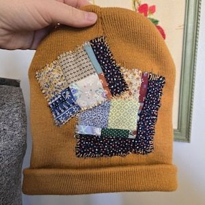 May include: Mustard yellow knit beanie with two patches of colorful fabric squares sewn on the front. The patches feature floral and patterned designs in blue, green, and red. The beanie is held by a hand.