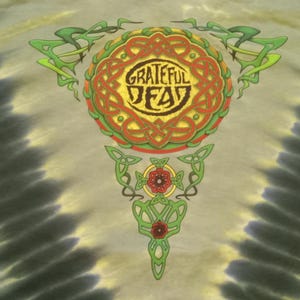 Puede incluir: Camiseta te&ntilde;ida con un dise&ntilde;o celta colorido, con las palabras "Grateful Dead" en un emblema circular. El dise&ntilde;o incluye elementos verdes, rojos y amarillos. El fondo tiene un patr&oacute;n de te&ntilde;ido anudado en tonos grises y amarillos.