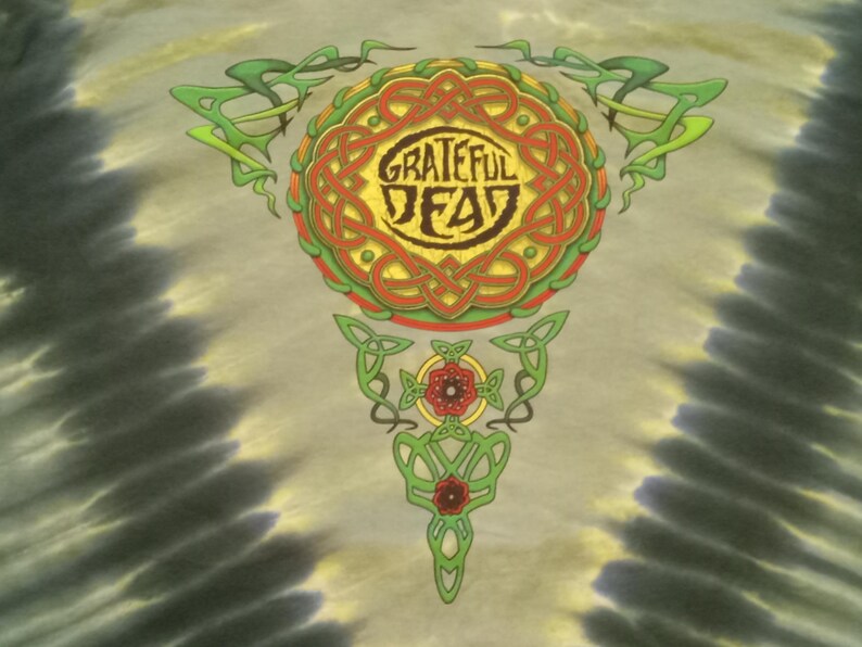 Puede incluir: Camiseta te&ntilde;ida con un dise&ntilde;o celta colorido, con las palabras "Grateful Dead" en un emblema circular. El dise&ntilde;o incluye elementos verdes, rojos y amarillos. El fondo tiene un patr&oacute;n de te&ntilde;ido anudado en tonos grises y amarillos.
