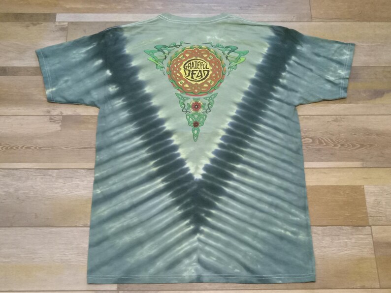 Puede incluir: Camiseta te&ntilde;ida con tinte anudado en tonos azules y verdes, con un dise&ntilde;o central con las palabras "Grateful Dead" en un patr&oacute;n circular. La parte posterior de la camiseta tiene un patr&oacute;n de te&ntilde;ido anudado en forma de V.