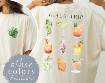 May include: Ivory-colored t-shirt with a tropical leaf and "Girls Trip" text. The back features watercolor illustrations of various cocktails. The shirt is a relaxed fit, perfect for a casual outing.