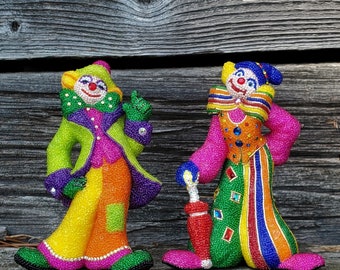 Puede incluir: Dos figuritas de payaso de cuentas de colores, una sosteniendo un paraguas rojo. Los payasos llevan ropa de color rosa brillante, verde, amarillo y naranja con rayas y patrones coloridos.