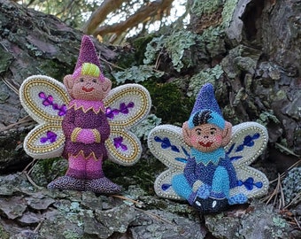 Puede incluir: Dos figuritas de hadas de cuentas con alas. El hada de la izquierda lleva un atuendo morado y rosa con alas doradas y moradas. El hada de la derecha lleva un atuendo azul con alas plateadas y azules. Ambas hadas están sentadas en un tronco de árbol.