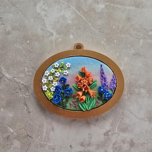 May include: Oval-shaped, gold-framed floral art piece. The artwork features a vibrant garden scene with blue, orange, and purple flowers, set against a blue sky background. The piece has a small loop at the top for hanging.
