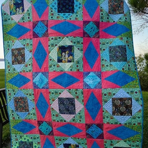 May include: A colorful handmade quilt with a geometric pattern. The quilt features a combination of teal, pink, and blue diamond and square shapes. The quilt is displayed outdoors, with a view of the sky and water in the background.