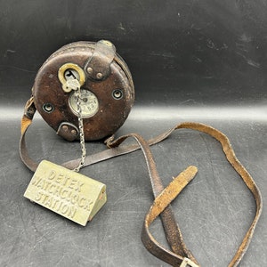 May include: A vintage leather watch clock with a metal chain and a key. The clock has a white face with black numbers and hands. The clock is attached to a brown leather strap with a metal buckle. The strap has a tag that reads "DETEX WATCHCLOCK STATION".