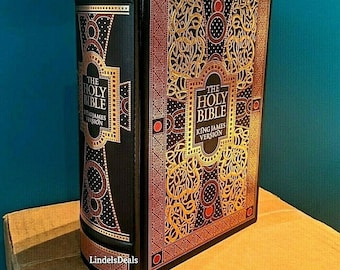 May include: A hardcover King James Version of The Holy Bible. The book has a black cover with gold and red decorative accents. The spine and cover feature the title in gold lettering. The book is on a cardboard surface.