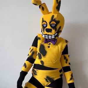 Op de afbeelding: Een geel en zwart Spring Bonnie kostuum. Het kostuum heeft een konijnenkop met zwarte markeringen, een paarse vlinderdas en een gele body met zwarte vlekken. De armen en benen zijn zwart. Het kostuum is ontworpen om op een personage uit de Five Nights at Freddy's-videogameserie te lijken.