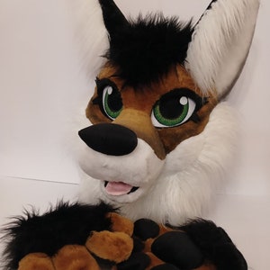 May include: A plush wolf head with black and white fur, brown and black paws, and green eyes. The wolf is smiling with its tongue sticking out.
