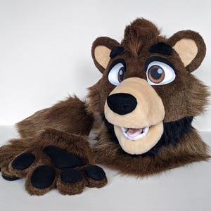 Op de afbeelding: Een bruine beren-fursuit hoofd- en pootset. De kop heeft grote, expressieve ogen, een beige snuit en een zwarte neus. De poten hebben zwarte voetzolen. De vacht is een mix van bruine tinten.