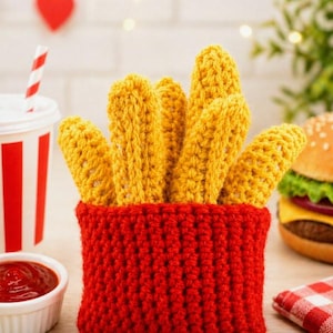 Könnte beinhalten: Ein roter gehäkelter Behälter gefüllt mit gelben gehäkelten Pommes Frites. Links befindet sich ein weißer Becher mit roten Streifen und einem rot-weiß gestreiften Strohhalm. Ein Burger und Ketchup sind im Hintergrund.