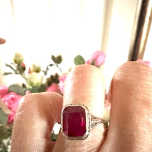 Op de afbeelding: Een zilveren ring met een grote, rechthoekige, dieprode edelsteen. De ring heeft een sierlijk filigraanontwerp rond de edelsteen en op de band. De achtergrond is wazig, met hints van roze bloemen en een wit gordijn.