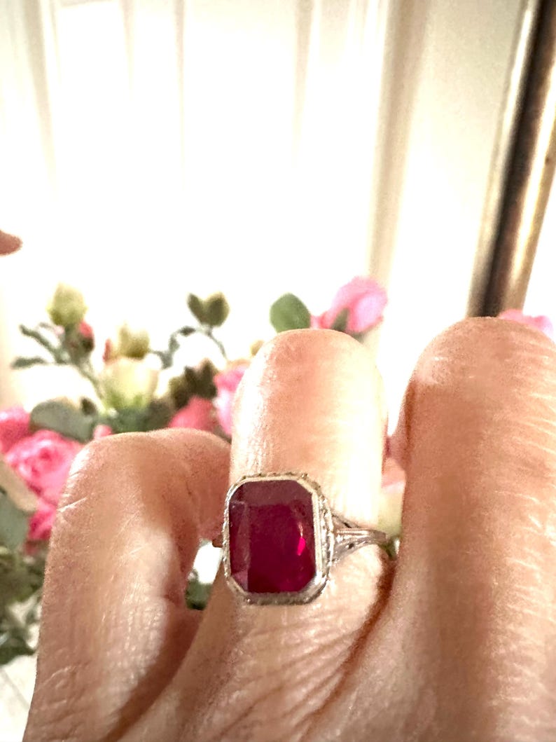 Op de afbeelding: Een zilveren ring met een grote, rechthoekige, dieprode edelsteen. De ring heeft een sierlijk filigraanontwerp rond de edelsteen en op de band. De achtergrond is wazig, met hints van roze bloemen en een wit gordijn.
