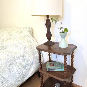 May include: A dark brown, three-tiered side table with a lamp and a vase. The lamp has a cream-colored lampshade and a geometric base. A light blue vase holds greenery. Books and a small container sit on the table.