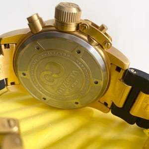 May include: Close-up of a gold-toned Invicta chronograph watch. The watch features a gold bezel, crown, and pushers. The watch back displays the Invicta logo and text. The watch band is black and gold. The watch is water resistant to 200 meters.