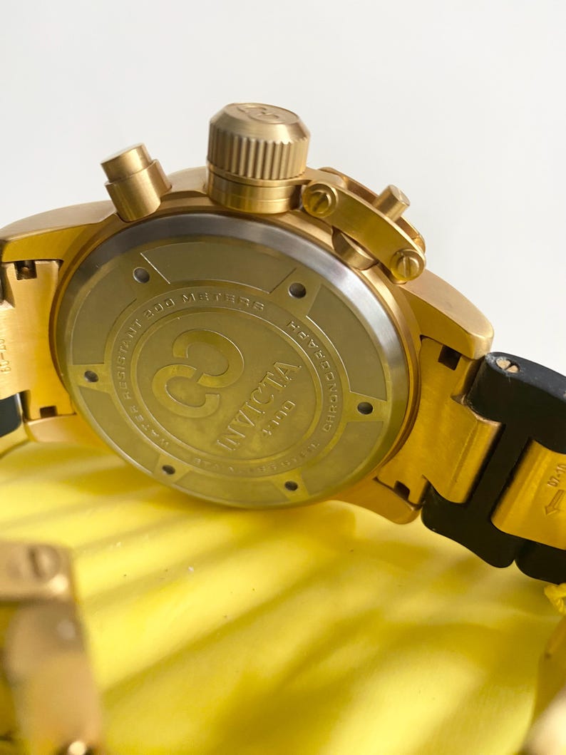 May include: Close-up of a gold-toned Invicta chronograph watch. The watch features a gold bezel, crown, and pushers. The watch back displays the Invicta logo and text. The watch band is black and gold. The watch is water resistant to 200 meters.