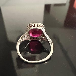 Op de afbeelding: Een zilveren ring met een grote, ovale, magenta gekleurde edelsteen. De ring heeft een sierlijk filigraanontwerp rond de edelsteen en op de band. De ring is geplaatst op een donkere, reflecterende ondergrond.
