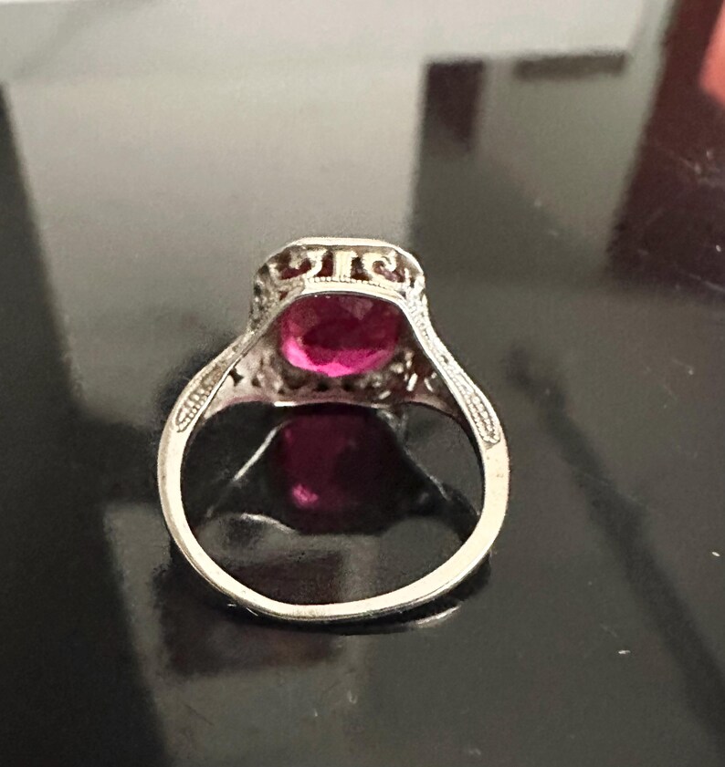 Op de afbeelding: Een zilveren ring met een grote, ovale, magenta gekleurde edelsteen. De ring heeft een sierlijk filigraanontwerp rond de edelsteen en op de band. De ring is geplaatst op een donkere, reflecterende ondergrond.