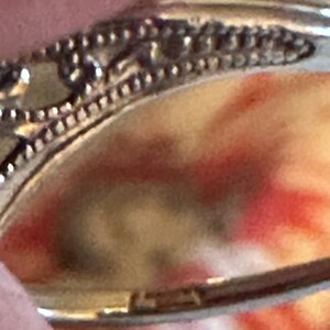 Op de afbeelding: Close-up van een zilveren ring met ingewikkelde filigraan details. De ring heeft een gladde band en de zetting heeft een decoratief patroon. De achtergrond is een vage mix van rood, oranje en geel.