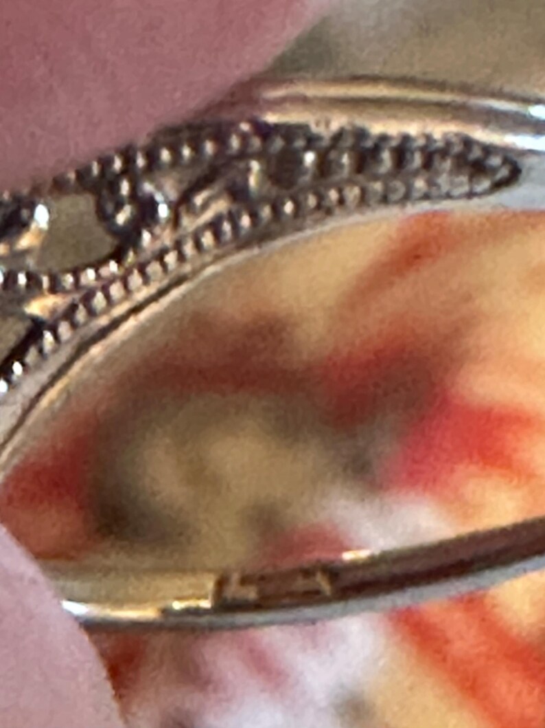 Op de afbeelding: Close-up van een zilveren ring met ingewikkelde filigraan details. De ring heeft een gladde band en de zetting heeft een decoratief patroon. De achtergrond is een vage mix van rood, oranje en geel.