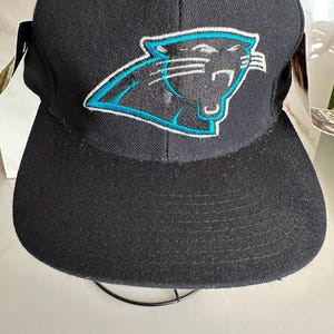 May include: Black baseball cap with the Carolina Panthers logo. The logo is a panther head in black and teal, with a white outline. The cap has a structured crown and a curved peak. The cap is made of a durable fabric.