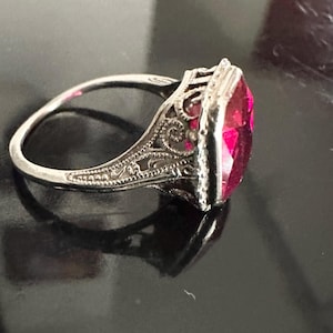 Op de afbeelding: Een zilveren ring met een sierlijk filigraanontwerp en een grote, rechthoekige, helder roze edelsteen. De ringband is dun en delicaat, en de edelsteen is gezet in een gedetailleerde bezel.