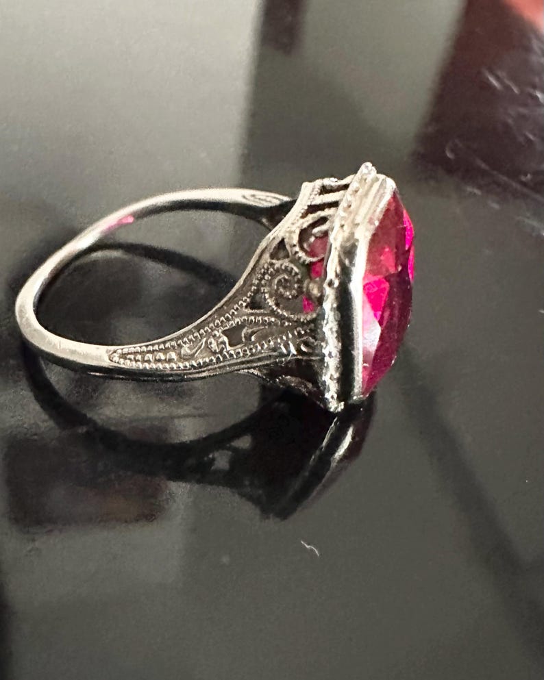 Op de afbeelding: Een zilveren ring met een sierlijk filigraanontwerp en een grote, rechthoekige, helder roze edelsteen. De ringband is dun en delicaat, en de edelsteen is gezet in een gedetailleerde bezel.