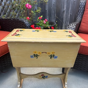 May include: A rectangular wooden sewing box with a light yellow finish, featuring hand-painted floral motifs in red, yellow, and blue on the lid, front panel, and lower support brace. The box has a hinged top and stands on four curved legs.