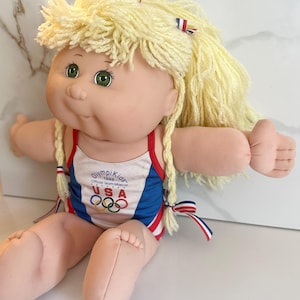 Könnte beinhalten: Cabbage Patch Kids Puppe mit blonden Garnhaaren und grünen Augen. Die Puppe trägt einen Badeanzug mit roten und blauen Verzierungen, die olympischen Ringe und den Text "Olympics Kids 1996 Official Team Mascot USA".