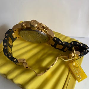 May include: Gold-toned wristwatch with a black and gold band. The watch features a round face and multiple dials. A yellow tag with the brand name is attached to the band. The watch is resting on a yellow surface.