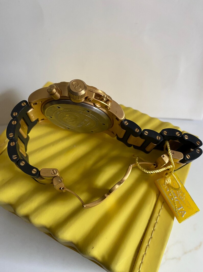 May include: Gold-toned wristwatch with a black and gold band. The watch features a round face and multiple dials. A yellow tag with the brand name is attached to the band. The watch is resting on a yellow surface.