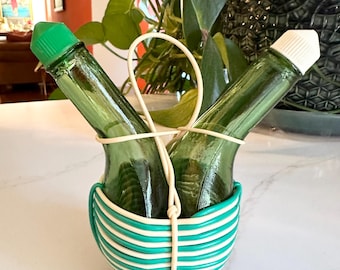 May include: Two green glass bottles with white tops are held together by a white and green striped wire holder. The bottles are in a criss-cross pattern.