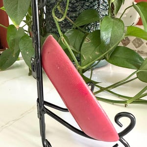 May include: A vibrant red object, possibly a decorative piece, is displayed on a black metal stand. The stand has a curved design with scroll-like details. The background features lush green plants, adding a natural contrast to the product.