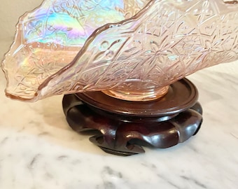 May include: A pink iridescent glass bowl with a scalloped edge and intricate floral pattern, displayed on a dark wood pedestal.