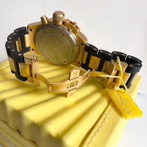 May include: Gold-toned Invicta chronograph watch with black accents on the band. The watch is displayed on a yellow surface, with a yellow tag attached. The watch features a round face and a clasp closure.