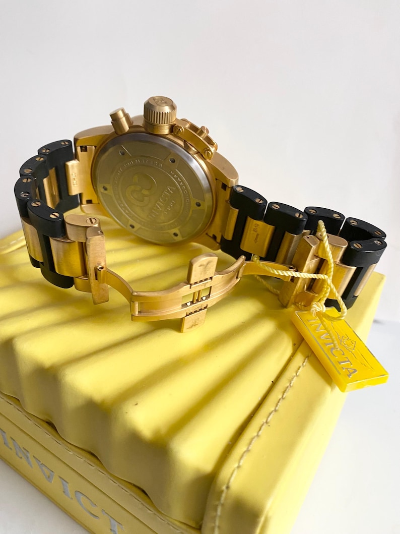 May include: Gold-toned Invicta chronograph watch with black accents on the band. The watch is displayed on a yellow surface, with a yellow tag attached. The watch features a round face and a clasp closure.