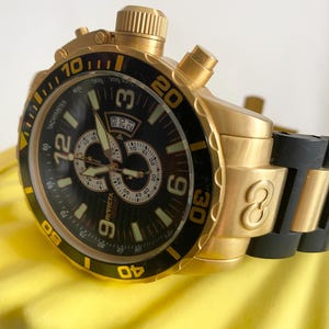 May include: A gold-toned Invicta chronograph watch with a black bezel and dial. The watch features a tachymeter, multiple sub-dials, and luminous hands. The band is black rubber. The watch has a date display at the 3 o'clock position.