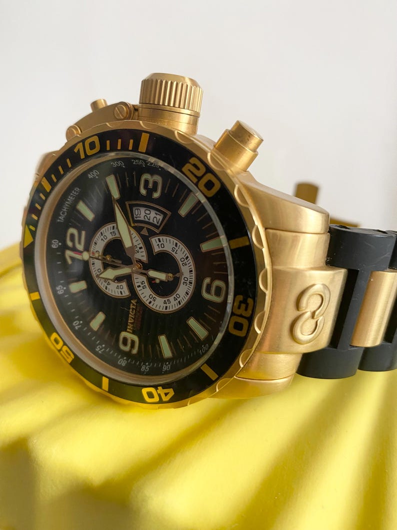 May include: A gold-toned Invicta chronograph watch with a black bezel and dial. The watch features a tachymeter, multiple sub-dials, and luminous hands. The band is black rubber. The watch has a date display at the 3 o'clock position.