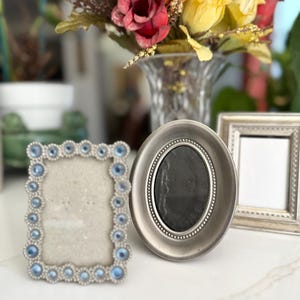 Peut inclure: Trois cadres photo argentés avec des designs différents. Le cadre de gauche a un design bleu clair et argent avec des strass. Le cadre du milieu est de forme ovale et a une finition argentée. Le cadre de droite est un simple cadre argenté avec un passe-partout blanc.