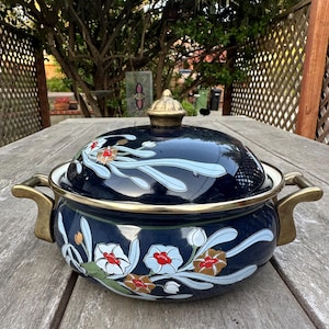 May include: A dark blue enamel casserole dish with a lid, featuring a floral design in white, red, and brown. The dish has gold-colored handles and trim. The lid is topped with a gold-colored finial. The dish is sitting on a wooden table.