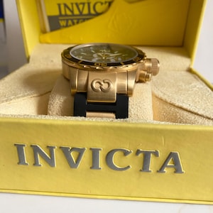 May include: A gold-toned Invicta watch with a black bezel and a black band, presented in a yellow box. The watch face is dark with gold accents. The box has the Invicta logo and text.