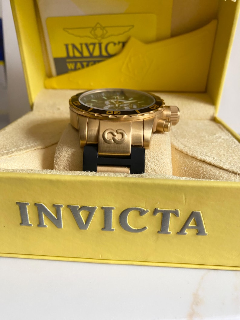 May include: A gold-toned Invicta watch with a black bezel and a black band, presented in a yellow box. The watch face is dark with gold accents. The box has the Invicta logo and text.